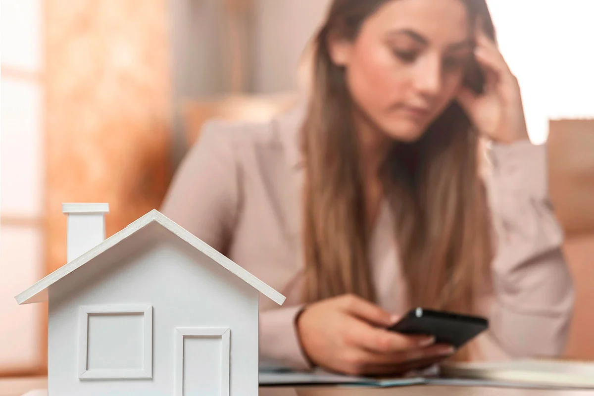 Mujer preocupada viendo su teléfono junto a una figura de casa, reflejando la pérdida de valor del dinero sin invertir en bienes raíces